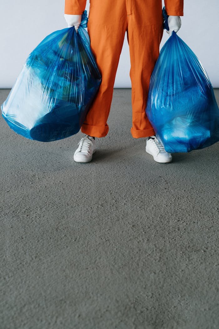 Person in orange uniform holding blue trash bags. Indoor setting focuses on waste management.